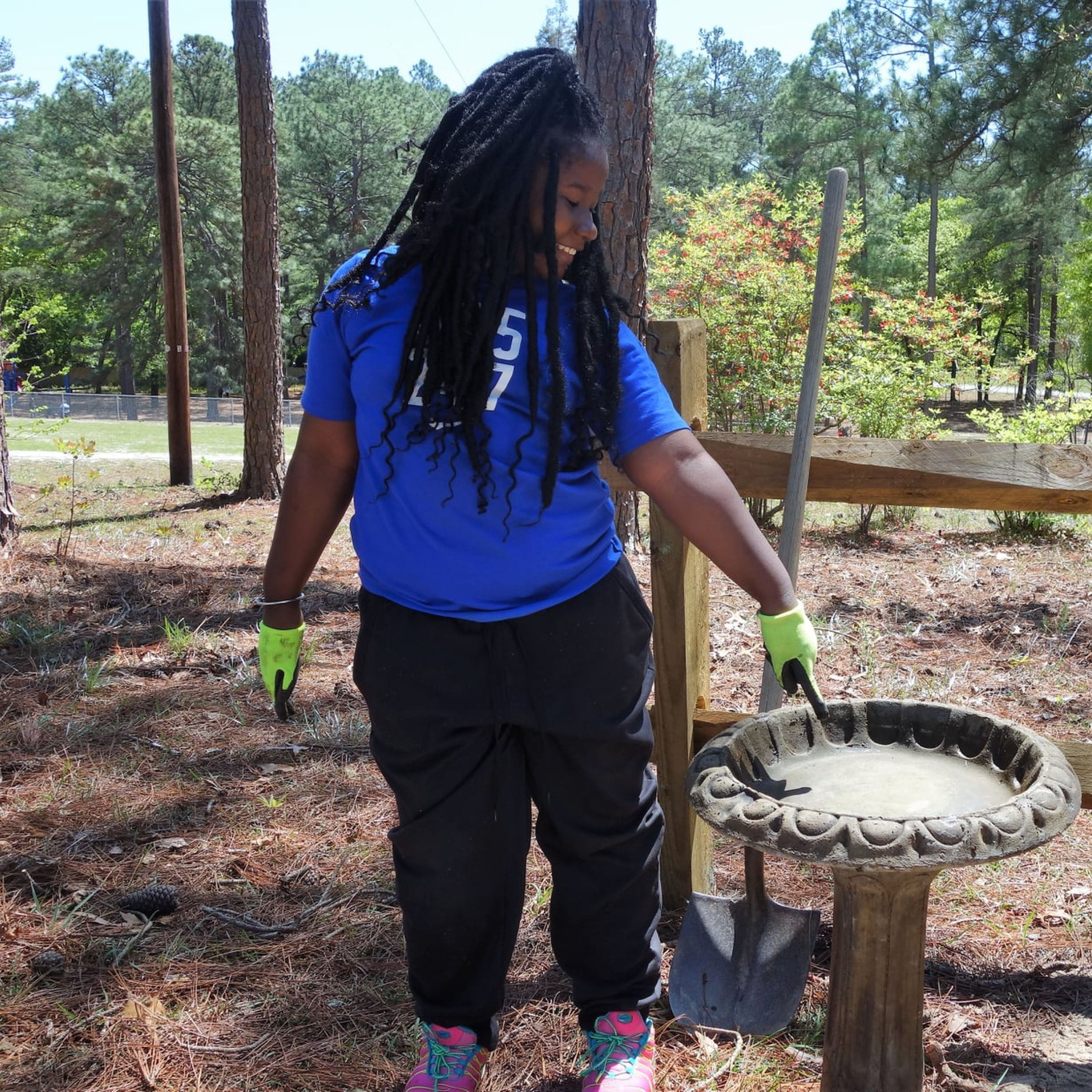 Young girl wearing work gloves, having worked in a school garden -- points to a bird bath.
