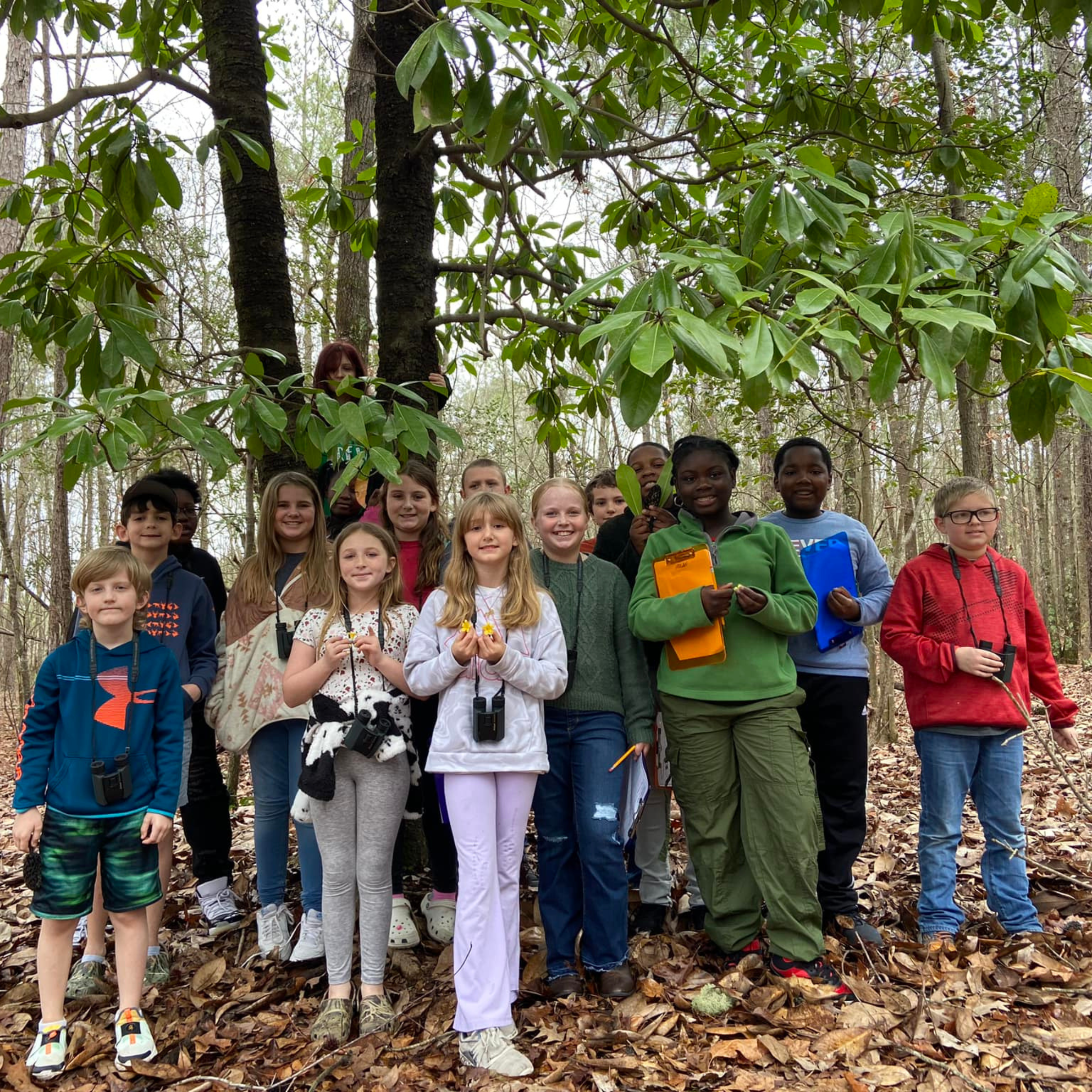 Group of middle school-aged children on a field trip in a forested landscape, holding clipboards and binoculars.